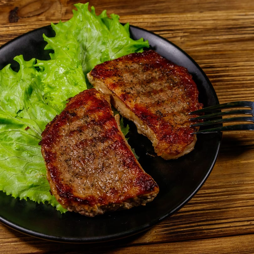 Two grilled pork chops with fresh lettuce on a black plate, with a fork on a wooden table.
