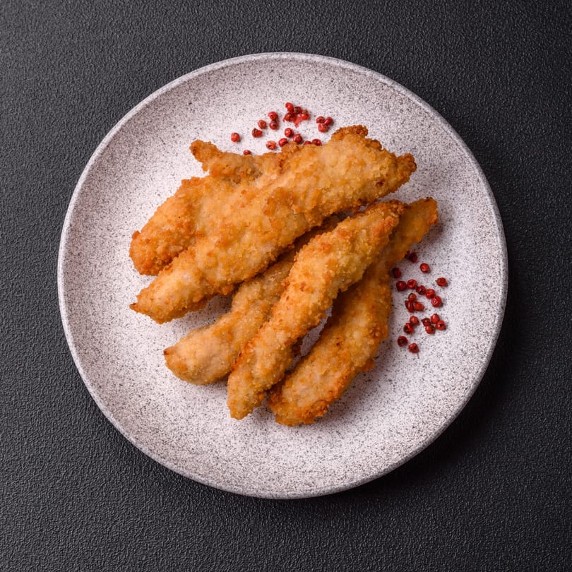 Crispy golden-brown chicken tenders with red peppercorns on a white speckled plate.