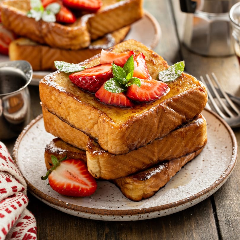 Stack of golden French toast with strawberries, powdered sugar, and mint on a rustic plate.