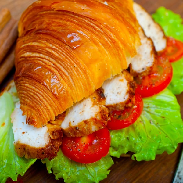 Golden croissant sandwich filled with grilled chicken, fresh lettuce, and sliced tomatoes on a wooden board.