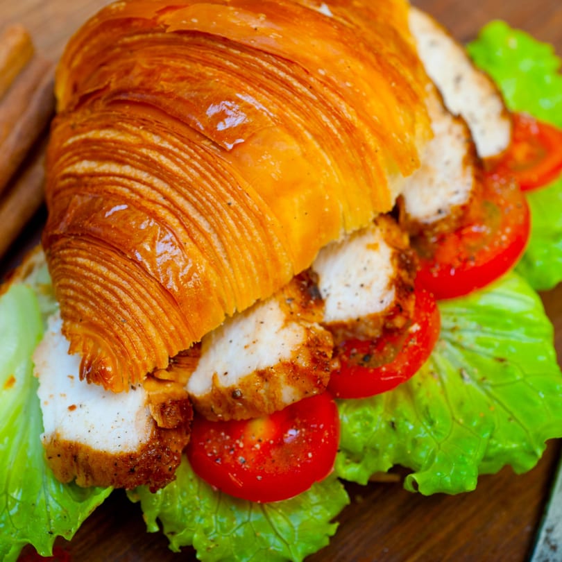 Golden croissant sandwich filled with grilled chicken, fresh lettuce, and sliced tomatoes on a wooden board.