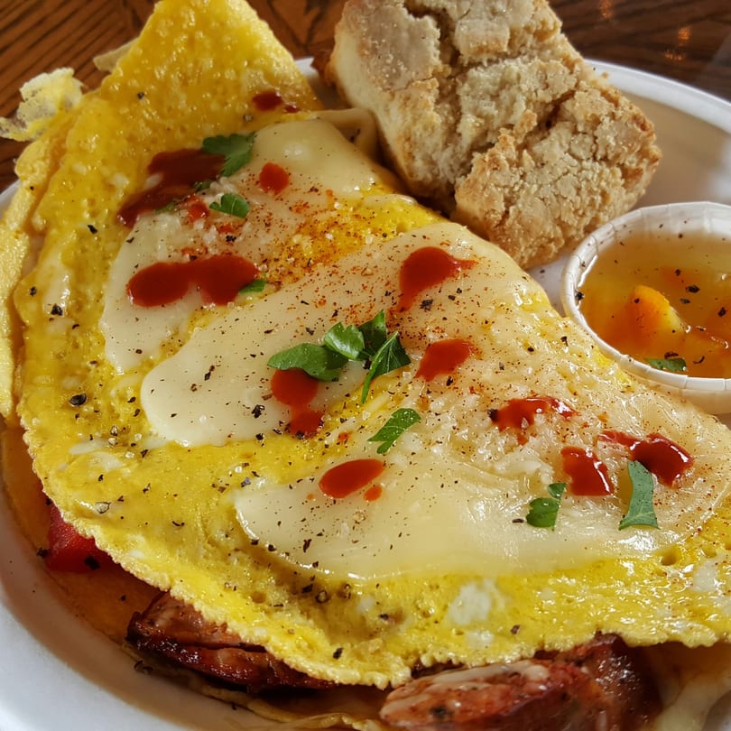 A breakfast omelet with melted cheese, hot sauce, and parsley, served with a biscuit and fruit preserves.