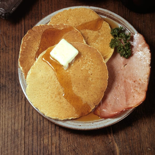 Pancakes with butter and syrup, served with a slice of ham and parsley on a plate.
