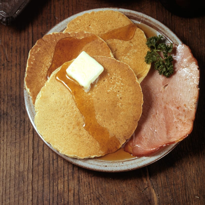 Pancakes with butter and syrup, served with a slice of ham and parsley on a plate.