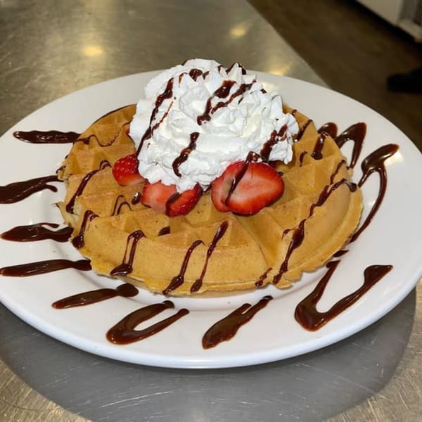 Golden waffle topped with whipped cream, sliced strawberries, and chocolate drizzle on a white plate.