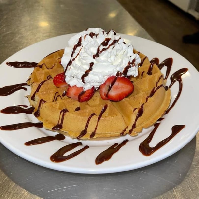 Golden waffle topped with whipped cream, sliced strawberries, and chocolate drizzle on a white plate.