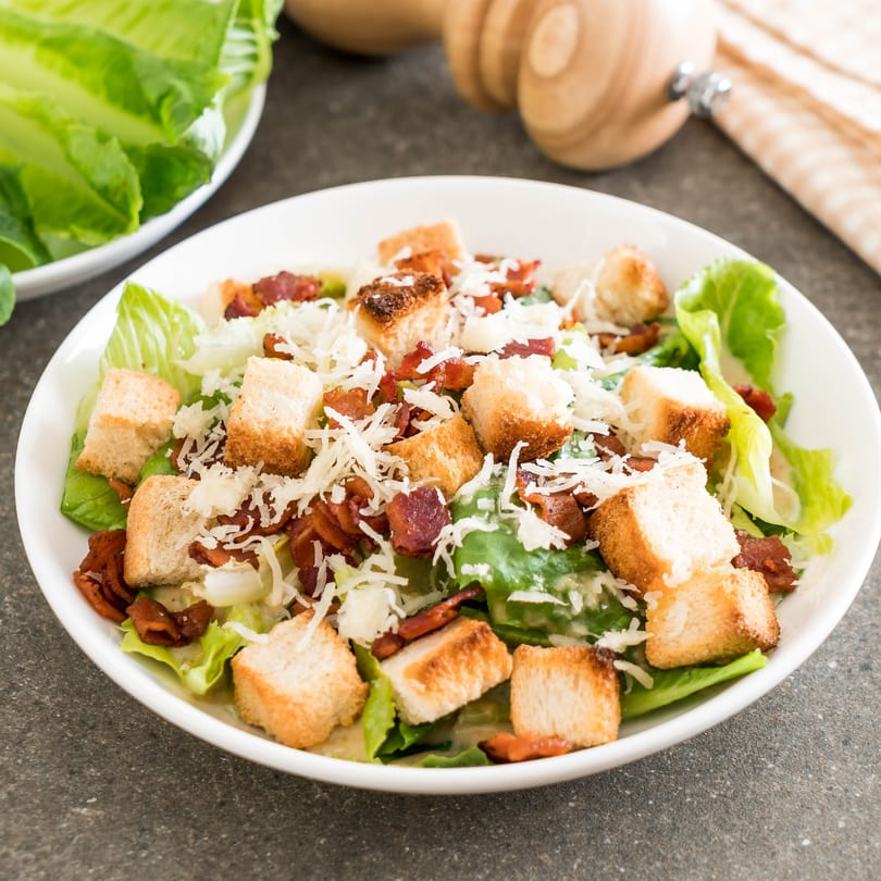 Caesar salad with crisp lettuce, bacon bits, croutons, and shredded cheese in a white bowl.